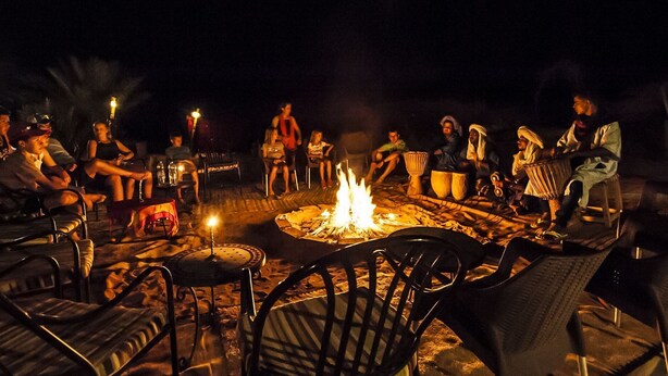 A group of people seated around a campfire at night, with chairs and tables arranged in a semi-circle.