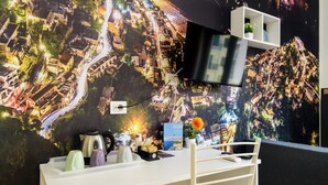 Une chambre d’hôtel moderne avec une grande fresque représentant un paysage urbain sur le mur, un téléviseur à écran plat, un bureau avec une bouilloire et une chaise.