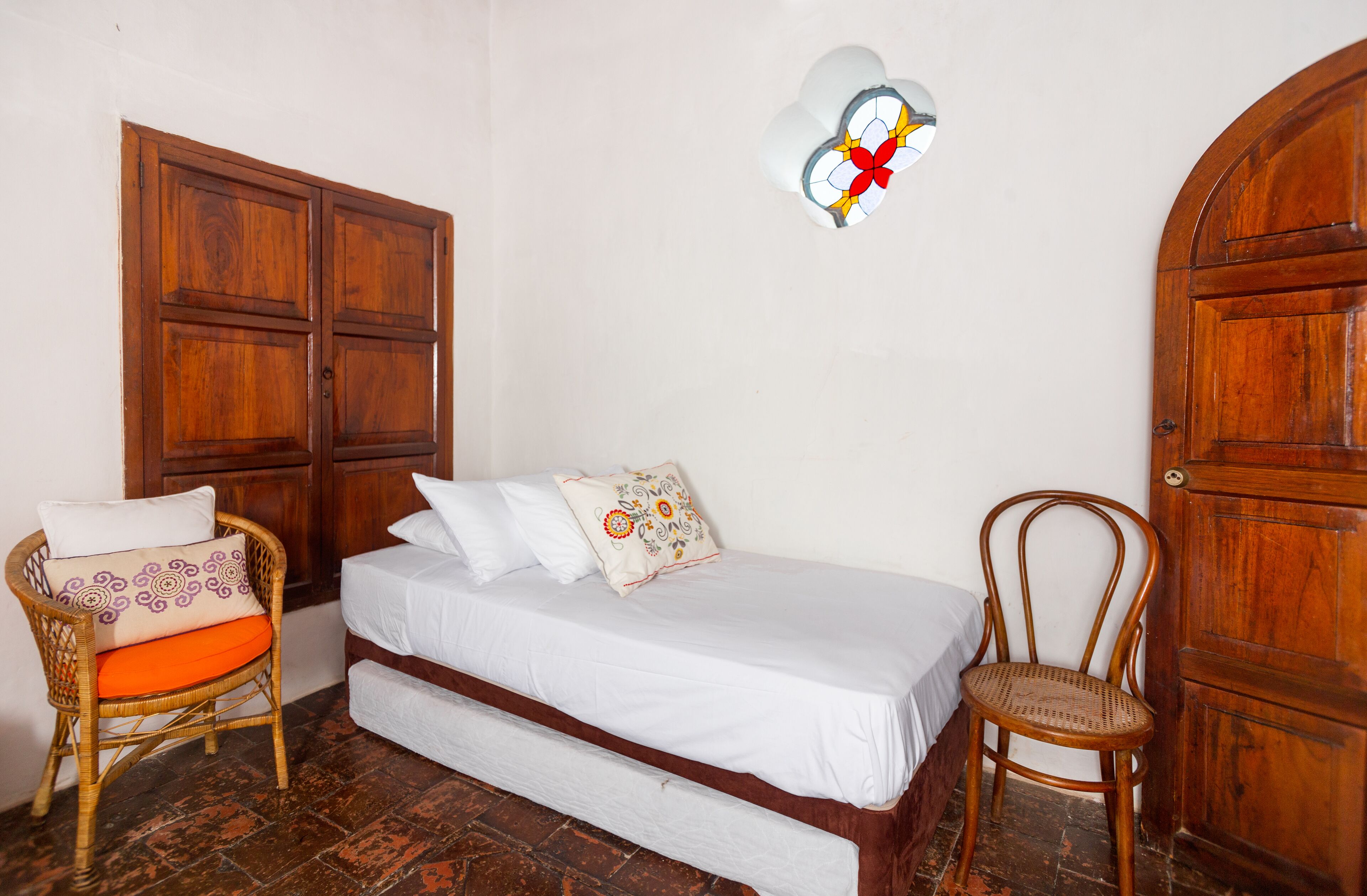 A room with a bed, a wicker chair, a wooden wardrobe, and a wall-mounted light fixture.