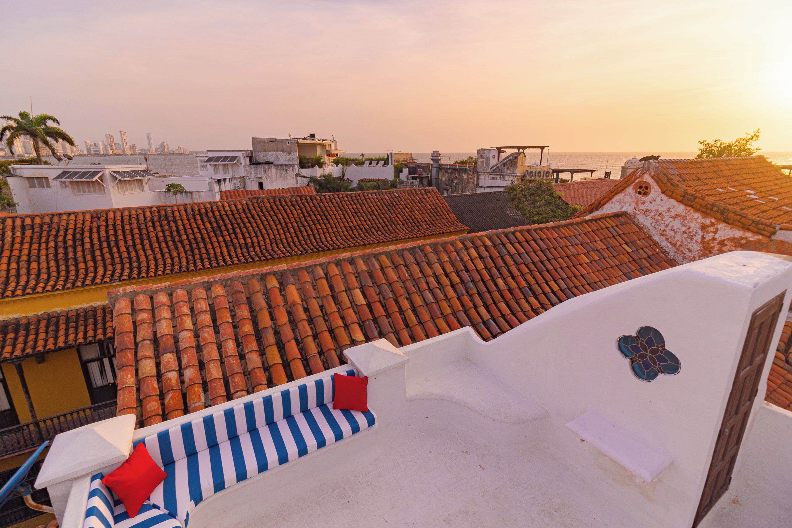 Terraza en la azotea con un sofá a rayas y un cojín rojo, con vistas a un paisaje urbano con tejados de terracota y una palmera.