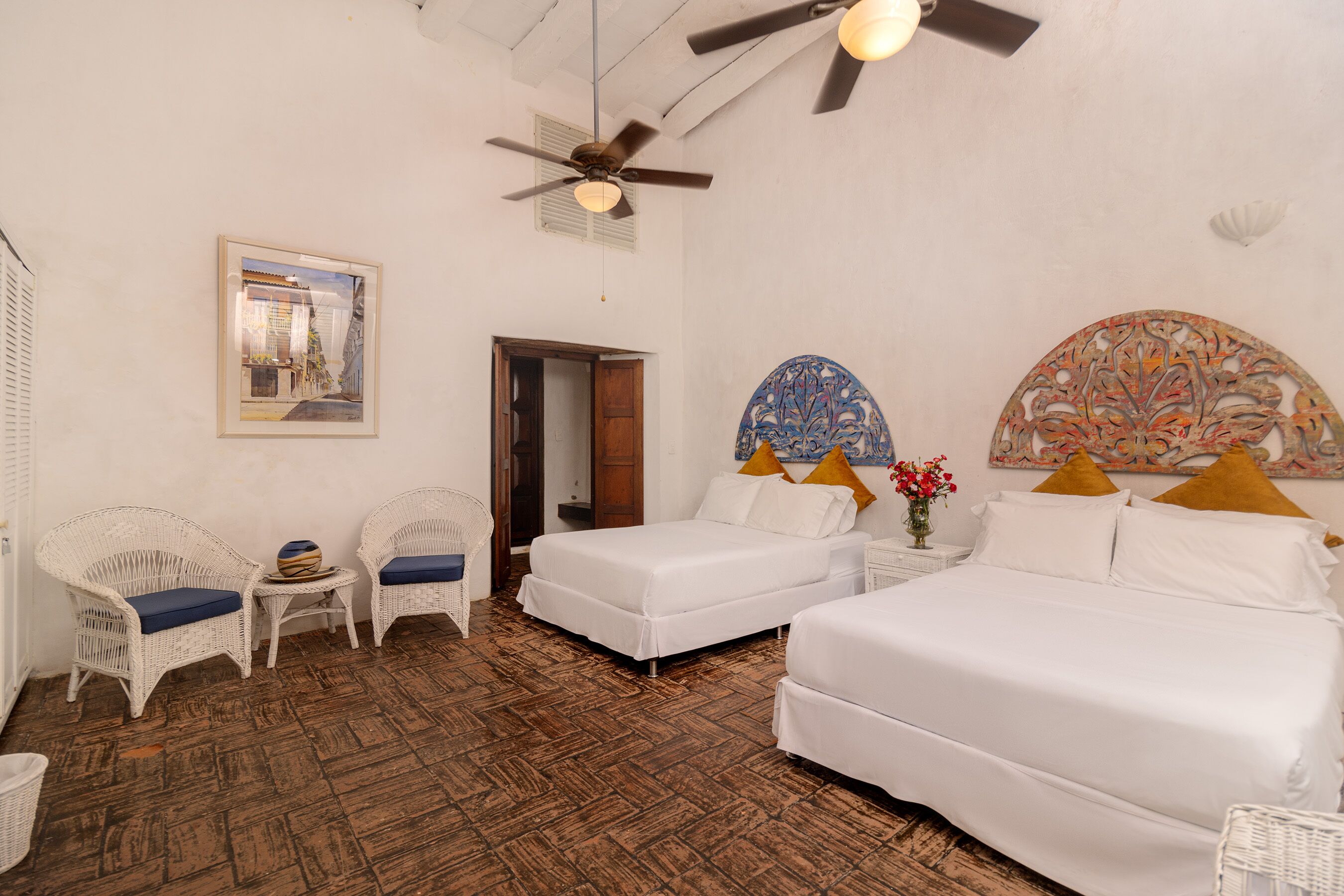 A hotel room with two beds, wicker chairs, a small table, and a ceiling fan.