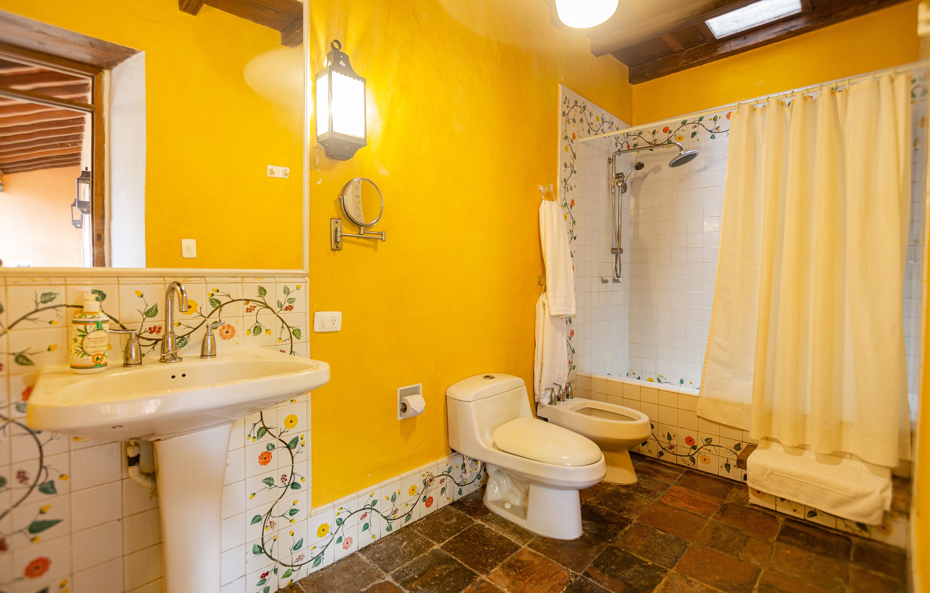 Un bany amb una paret groga, un lavabo, dues vàteres, una dutxa i un mirall.