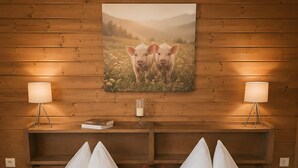 A hotel room with wooden panel walls, a painting of pigs, two bedside lamps, a bedside table with a book and a candle, two beds with white linens and red pillows, and rolled towels on the beds.