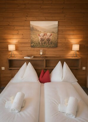 Camera d'albergo con pannelli in legno, due letti con lenzuola bianche, un cuscino rosso, lampade ai comodini, un quadro appeso alla parete e un piccolo tavolo con una candela.