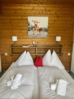 Hotellihuone, jonka seinät on verhoiltu puupaneeleilla, seinällä on lehmäaiheinen taulu, huoneessa on kaksi yöpöytää, joissa on lamput, ja sängyt on siististi pedattu pyyhkeineen.