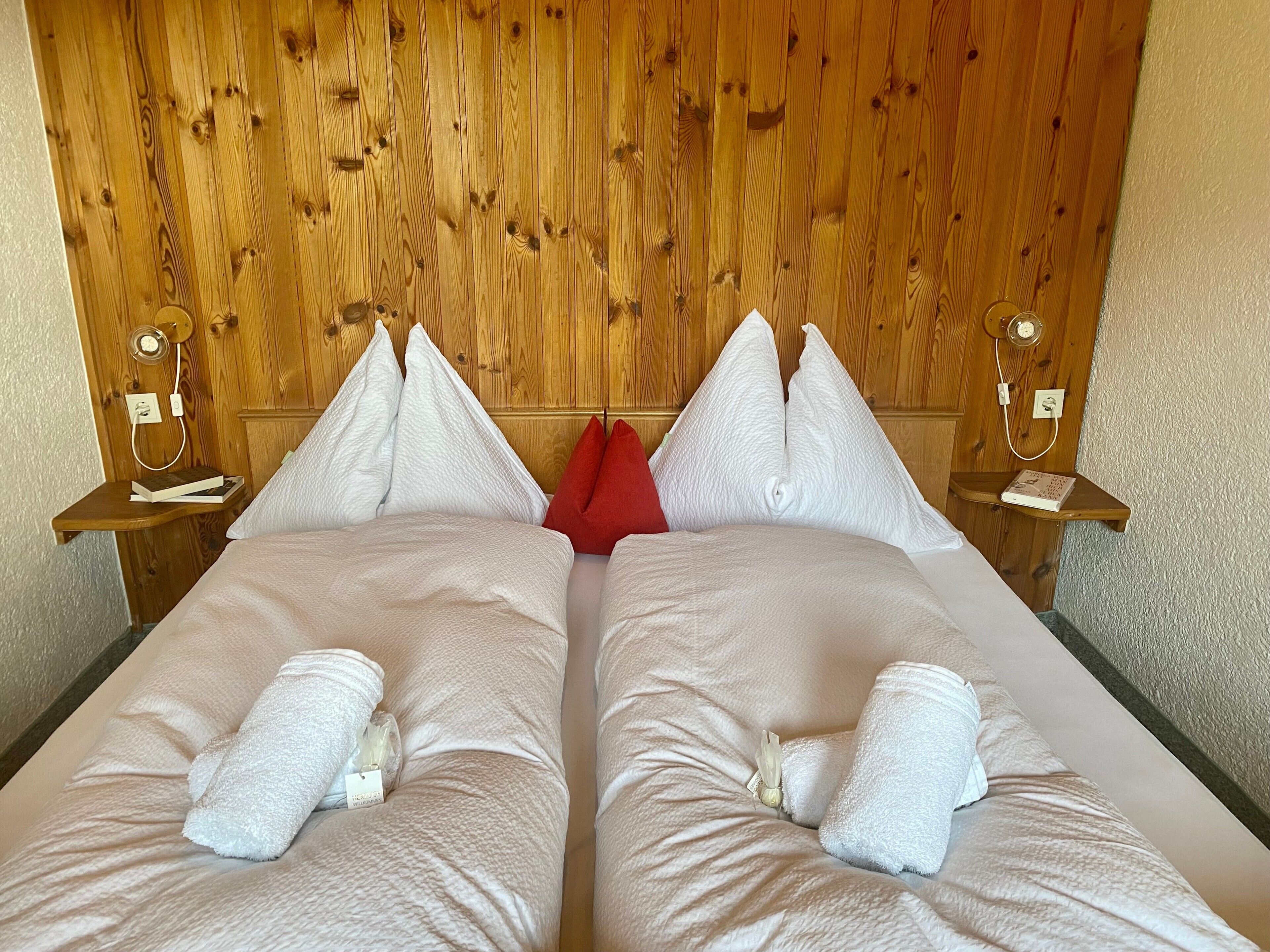 A double bed with white linens and pillows, wooden headboard, bedside tables with lamps, and a red accent pillow.