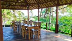 A covered outdoor dining area with a wooden table and chairs, surrounded by a wooden deck and a view of lush greenery.