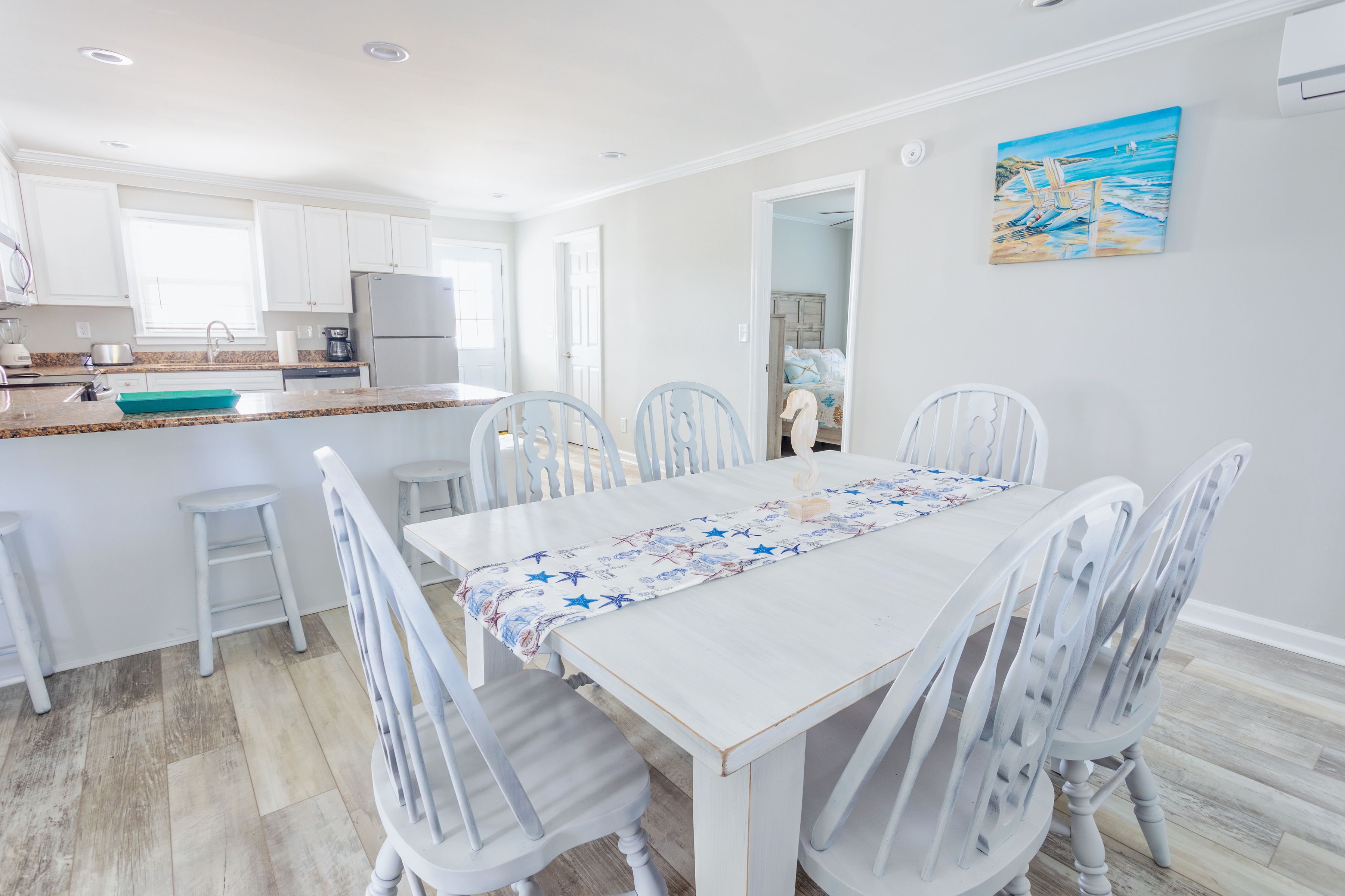 A dining area with a white table and chairs, a kitchen in the background, and a painting on the wall.