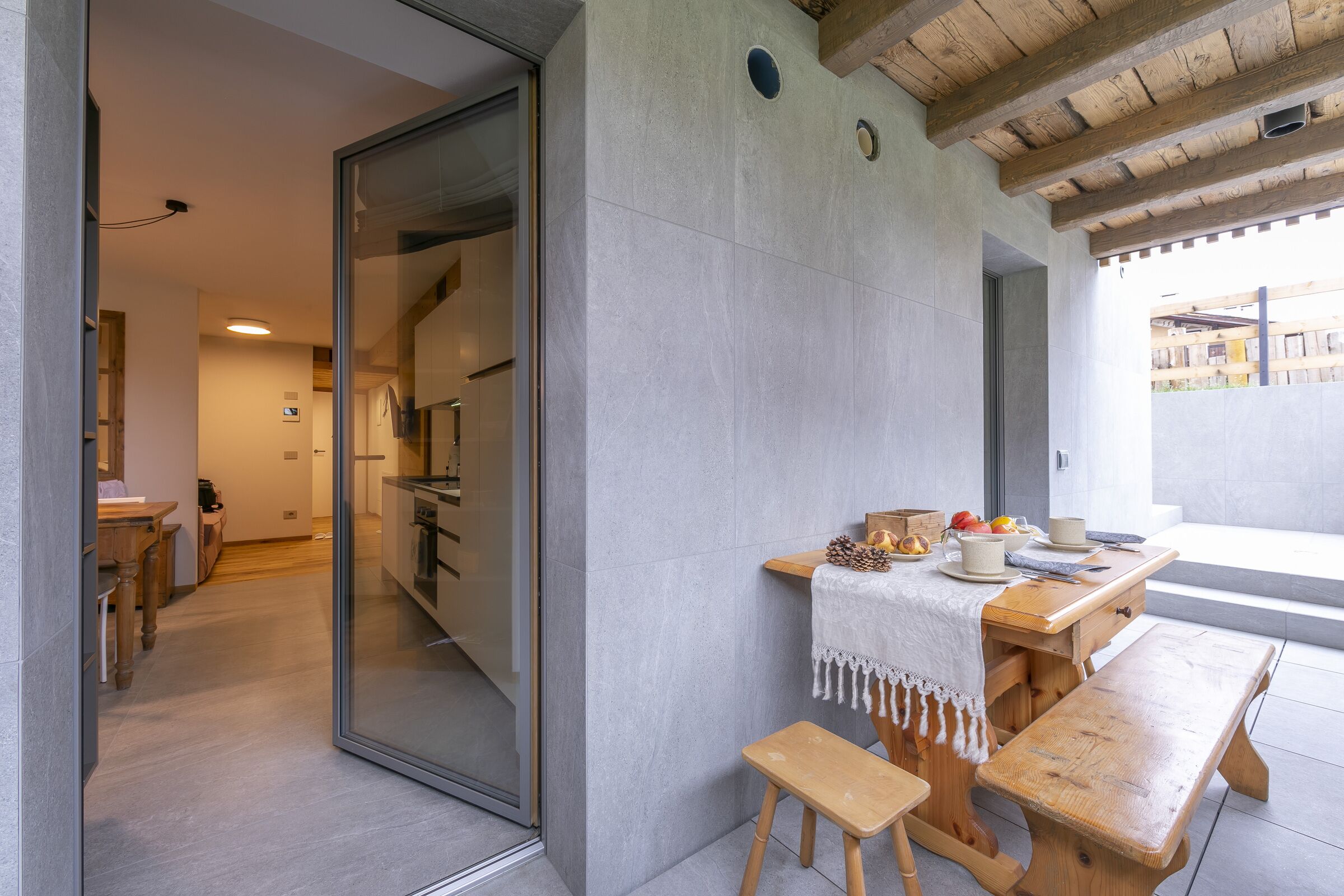 A modern kitchen with a wooden table and bench, a glass door leading to another room, and a view of the outside.