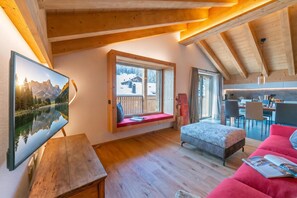 Un ambiente accogliente con soffitto in legno, un'ampia finestra con vista su un paesaggio innevato, una zona pranzo e un angolo salotto con un divano rosso.