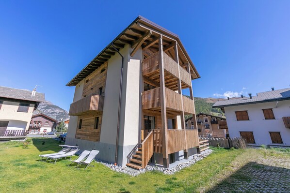 Un edificio de varios pisos con un balcón de madera, una escalera y un área verde con tumbonas.