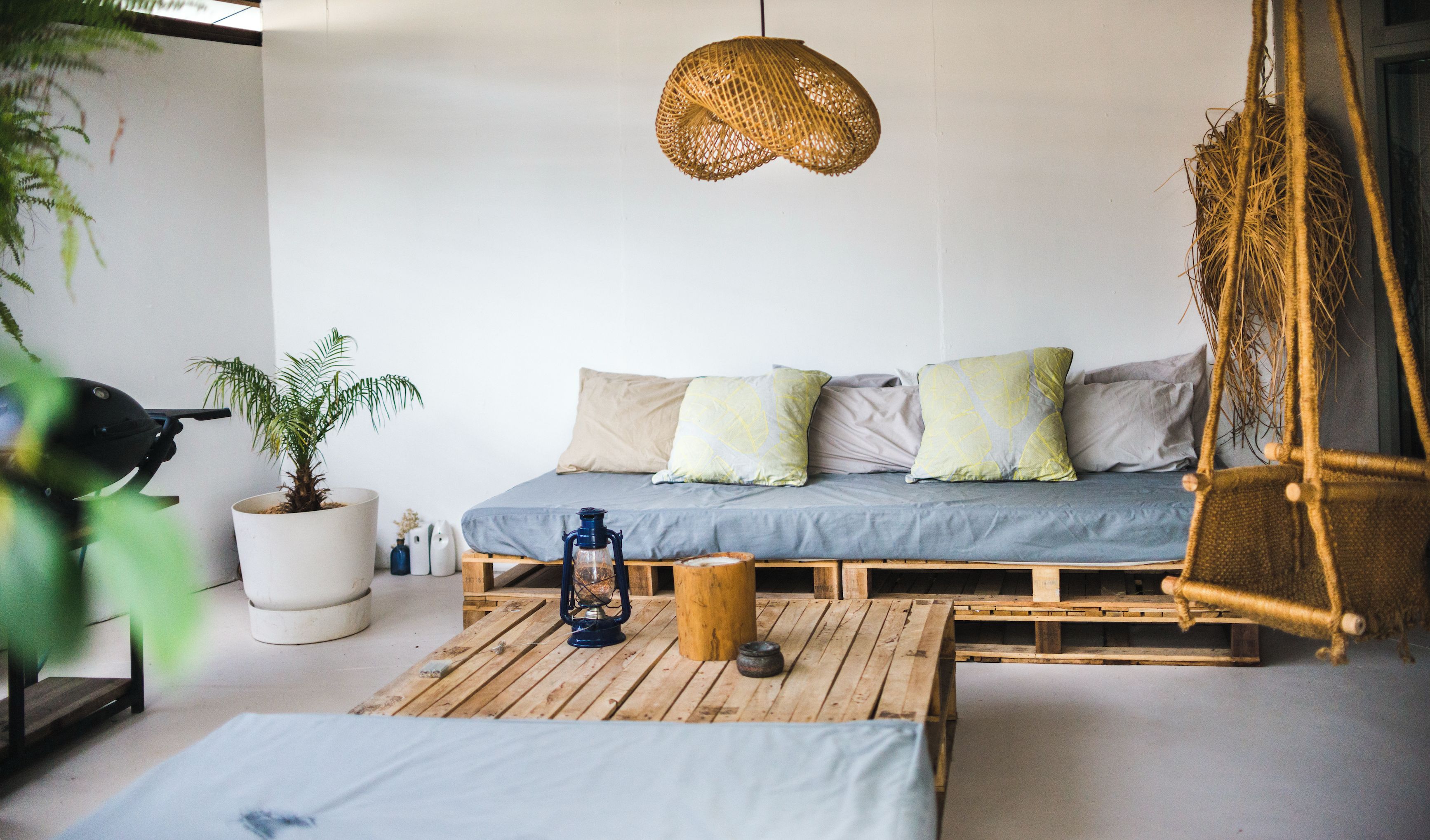 Ein gemütliches Zimmer mit einem Schlafsofa aus Holzpaletten, einem hängenden Rattan-Sessel, einer Topfpflanze und einer Laterne auf einem Holztisch.