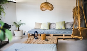 A cozy room with a wooden pallet sofa, a hanging rattan chair, a potted plant, and a lantern on a wooden table.