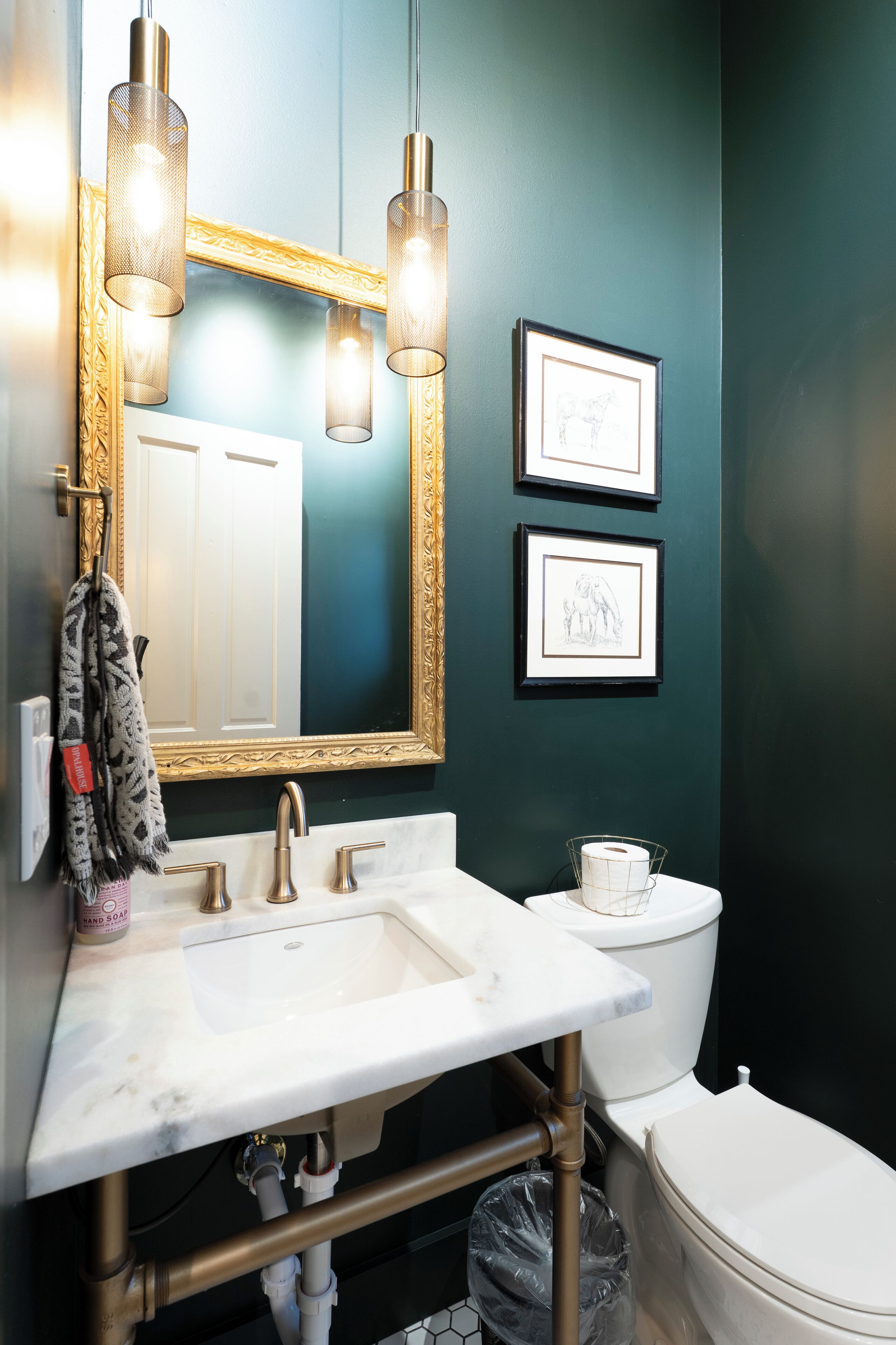 Ein Badezimmer mit einem weißen Waschbecken, goldenen Armaturen, einem spiegeln Rahmen aus Gold und zwei gerahmten Bildern an der Wand.