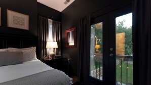 A bedroom with a bed, a nightstand, a lamp, a framed mirror, and a view of a balcony with a lawn and a fence.