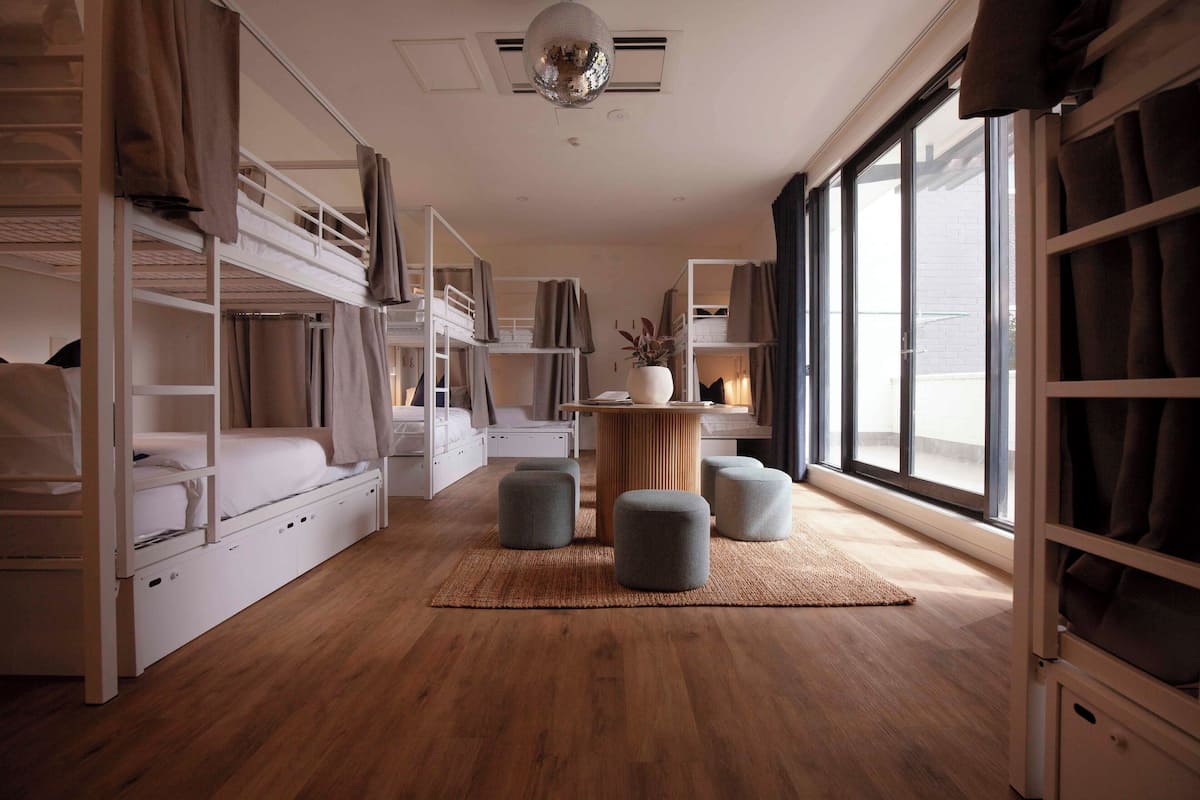 A modern, well-lit room with bunk beds, a central round table, and a large window providing natural light.