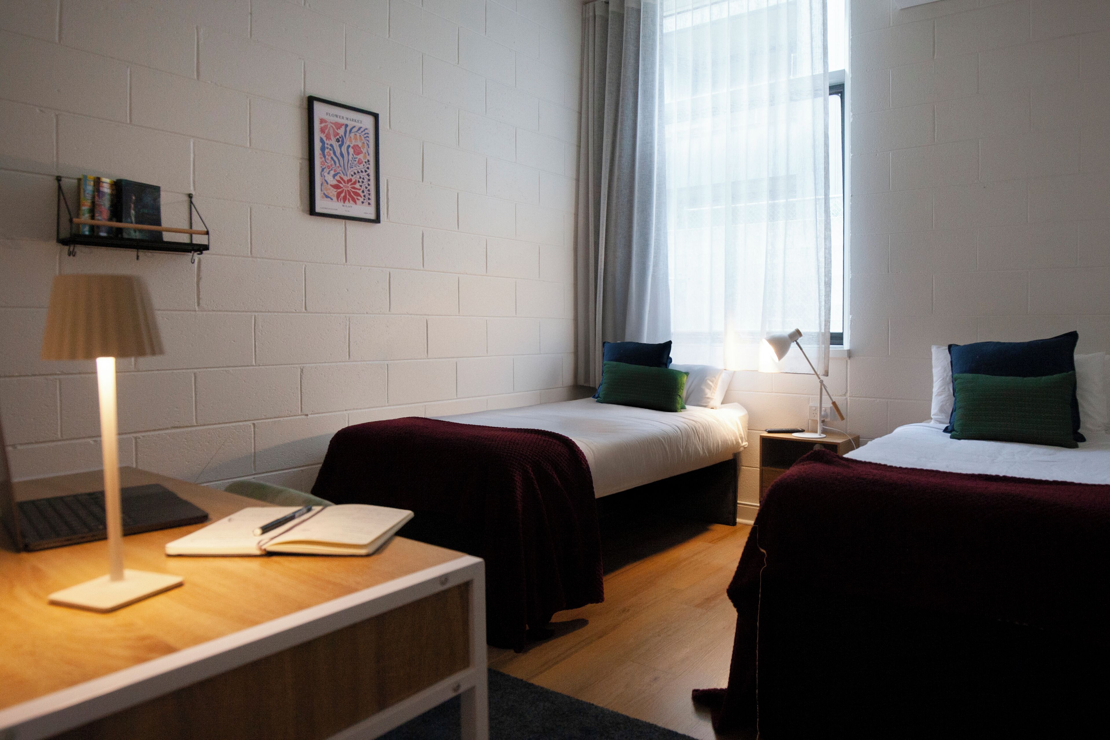 Huoneessa on kaksi sänkyä, työpöytä, jossa on kannettava tietokone ja muistikirja, lamppu, ikkuna, jossa on verhot, ja seinällä oleva kehystetty kuva.