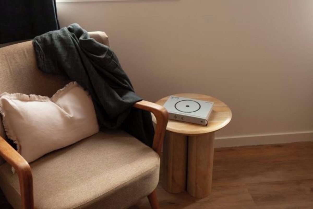 A chair with a cushion and a blanket, a small round wooden table with a device on it, and a plain wall.