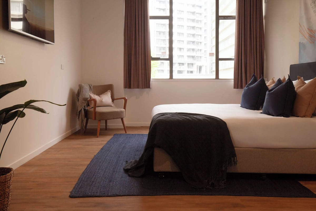 A bedroom with a bed, a chair, a window with curtains, and a plant.