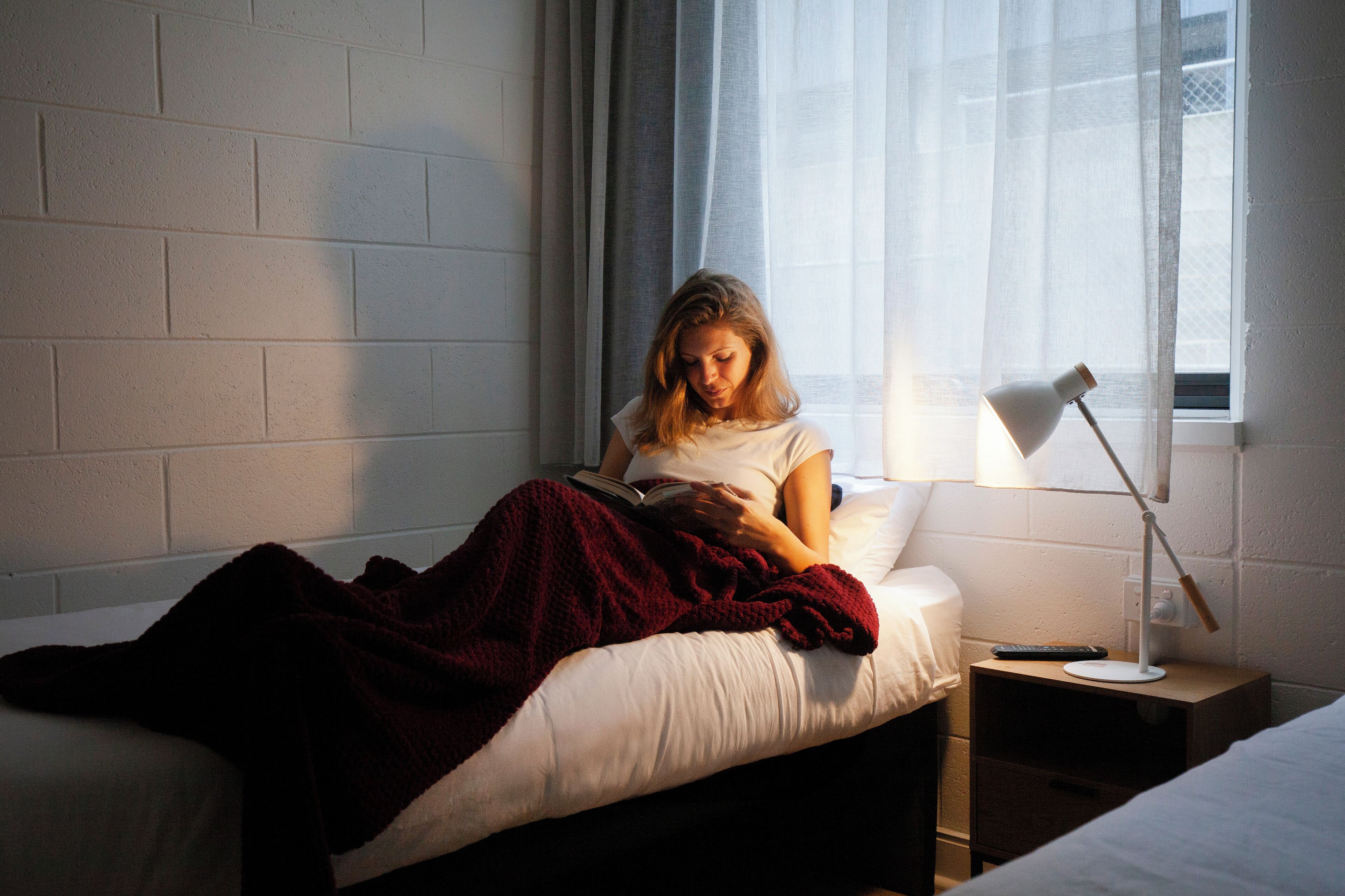 A person reading a book in a room with a white brick wall, a bedside table with a lamp, and a window with sheer curtains.