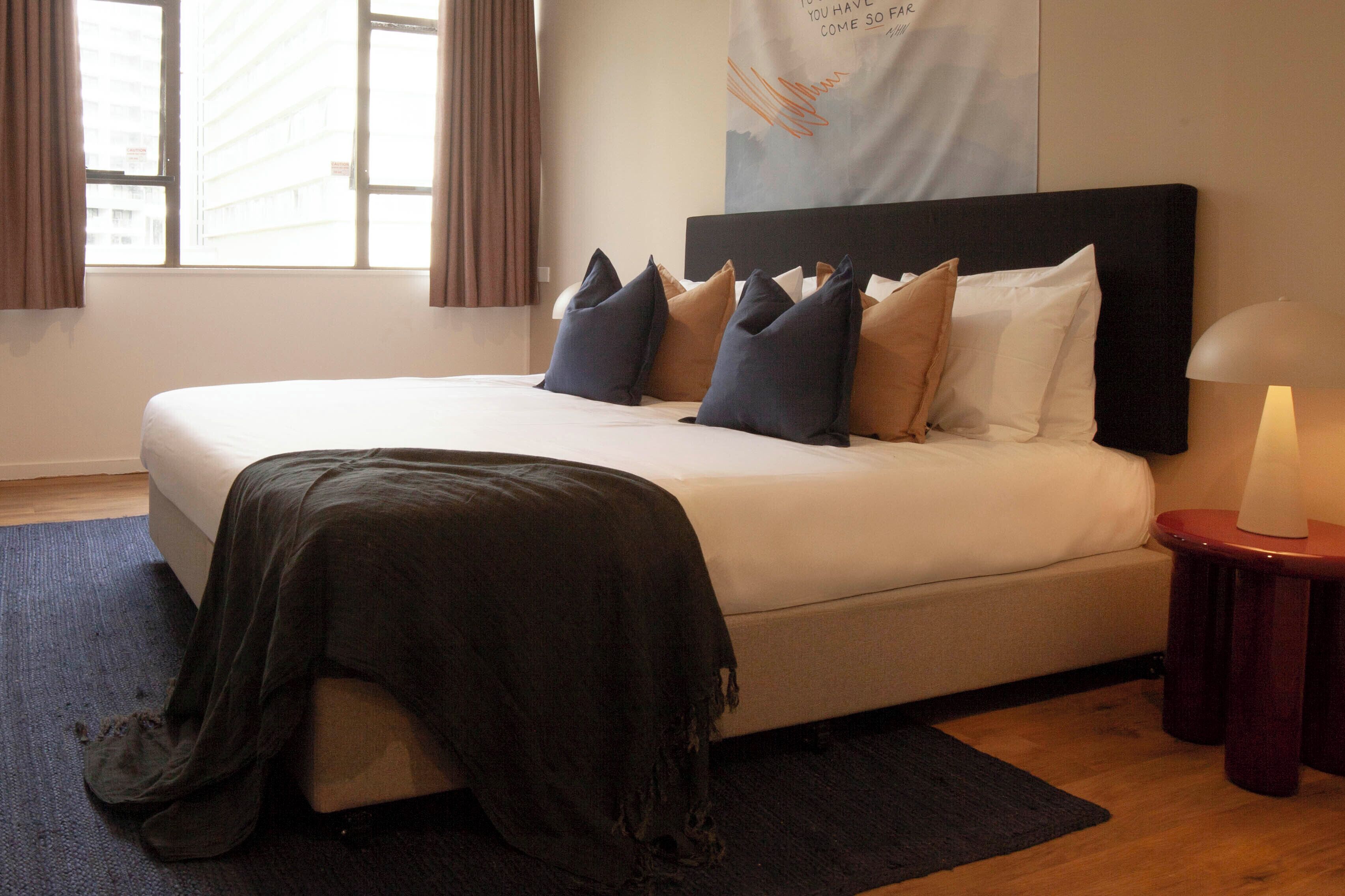 A neatly made bed with a black headboard, a dark throw blanket, and several pillows in shades of blue and beige.