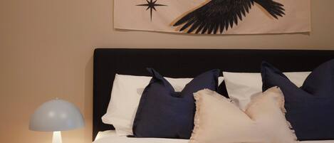 Una cama bien hecha con almohadas blancas y azul oscuro, una mesita de noche roja con una lámpara, y un cuadro en la pared sobre la cama.