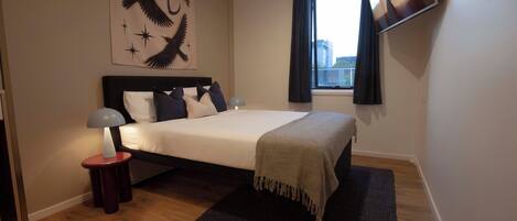 A bedroom with a bed, a nightstand, a lamp, a window with curtains, and a wall-mounted TV.