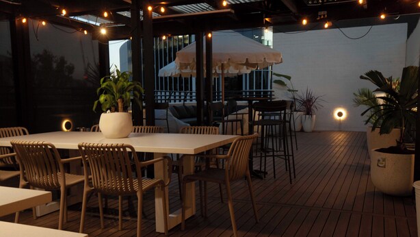 Un patio al aire libre con piso de madera, mesas, sillas, plantas en macetas y luces colgantes.