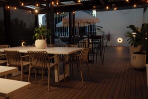 Un patio al aire libre con piso de madera, mesas, sillas, plantas en macetas y luces colgantes.