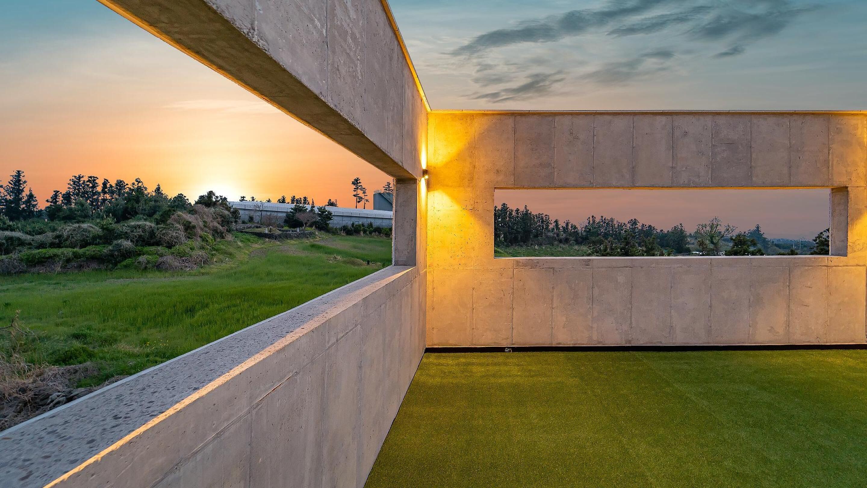 A modern architectural structure with a concrete exterior and a window providing a view of a sunset over a grassy landscape.