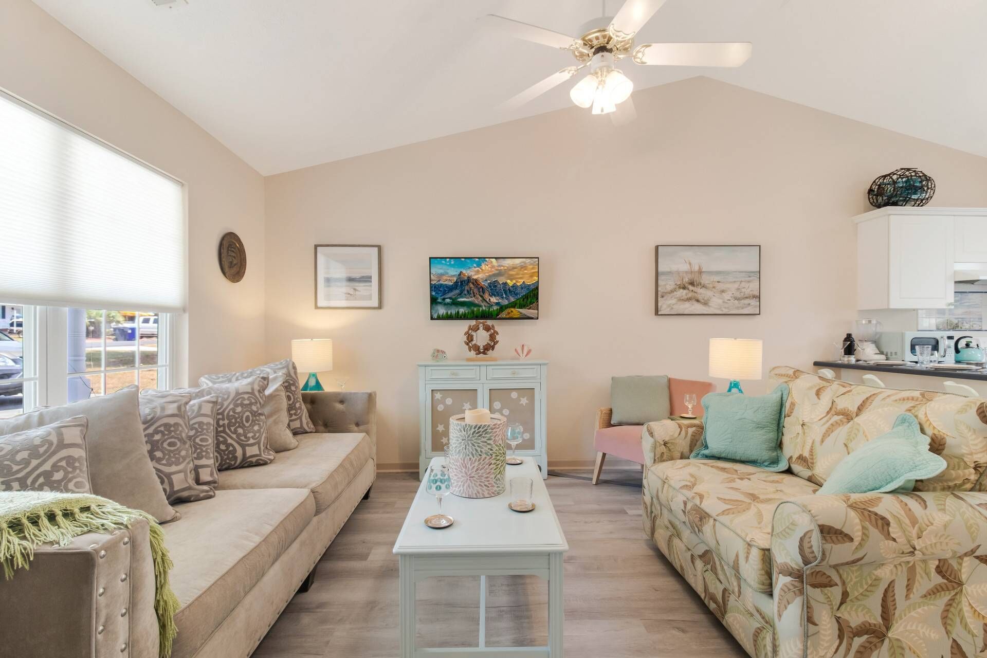A spacious living room with a high ceiling, a ceiling fan, and a large window. There are two sofas, a coffee table, and a dining table with chairs. The room has a neutral color palette and features artwork on the walls.