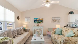 A spacious living room with a high ceiling, a ceiling fan, and a large window. There are two sofas, a coffee table, and a dining table with chairs. The room has a neutral color palette and features artwork on the walls. - Dog Friendly Cottage w/ Large Fenced in Backyard - Close to Beach (Oak Island)
