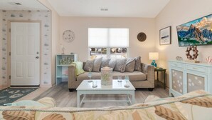A living room with a sofa, coffee table, and decorative items. There is a window with blinds, a door, and a wall with a painting. - Dog Friendly Cottage w/ Large Fenced in Backyard - Close to Beach (Oak Island)