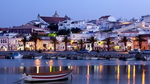 A coastal town at dusk with boats docked along the waterfront, buildings with balconies, and a prominent church spire. - Central Apartment Near the Beaches (Lagos)