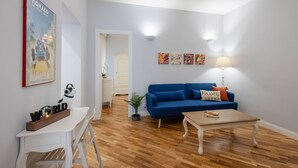 En stue med en blå sofa, et hvidt bord og en lampe. Der er et indrammet billede på væggen og en lille plante på gulvet.