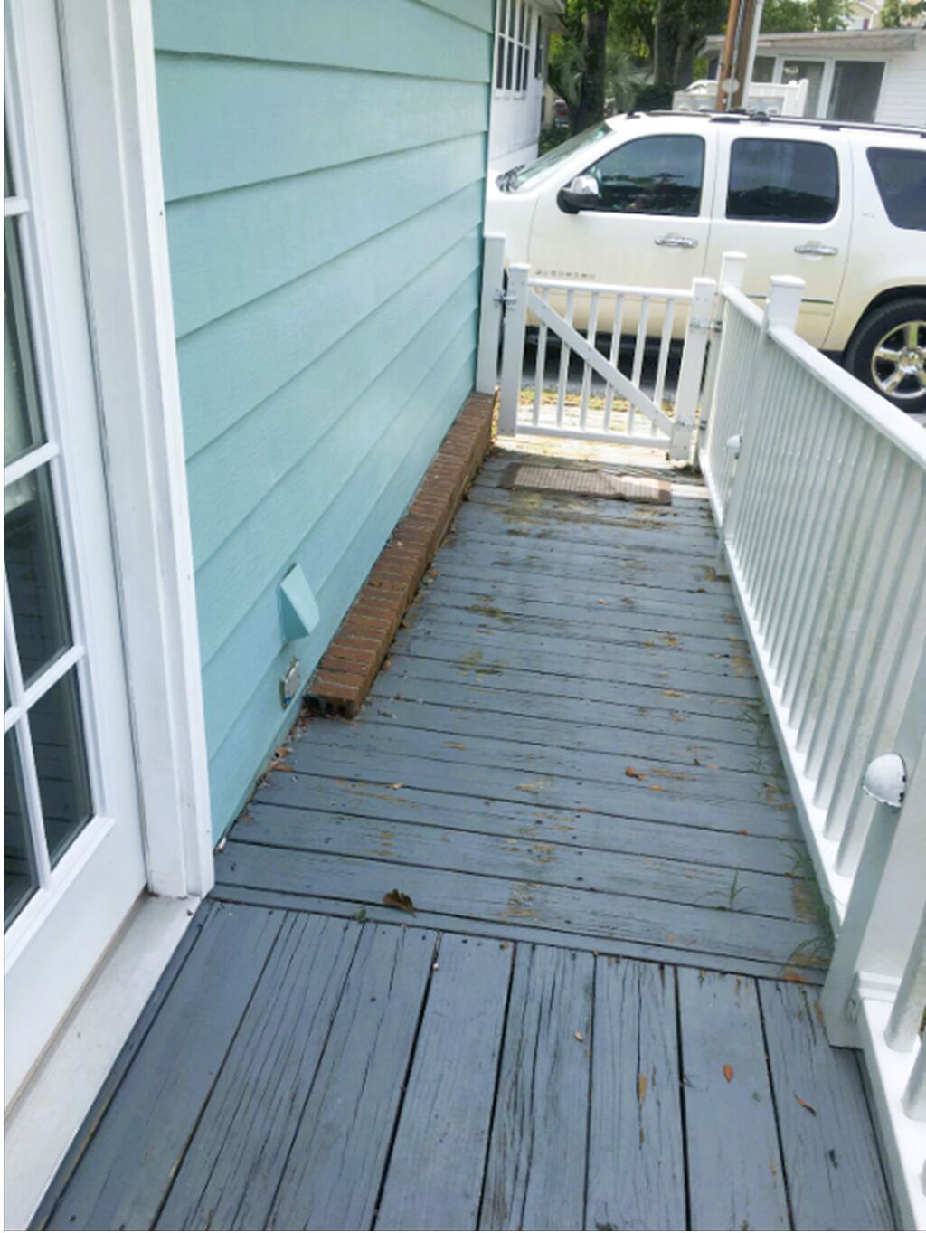 A wooden deck with a white railing, a white vehicle parked in the background, and a brick ledge at the bottom.