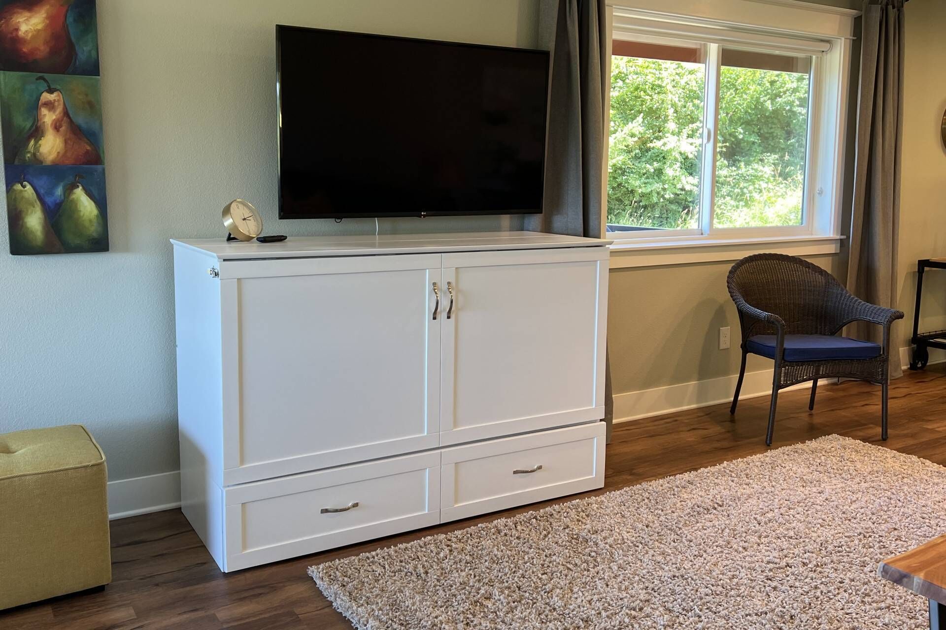 A room with a white cabinet, a flat-screen TV, a wicker chair, and a window with a view of trees.