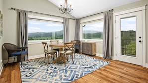 Un comedor con una mesa y sillas, un tapiz estampado, una araña y ventanas grandes con vistas al campo.