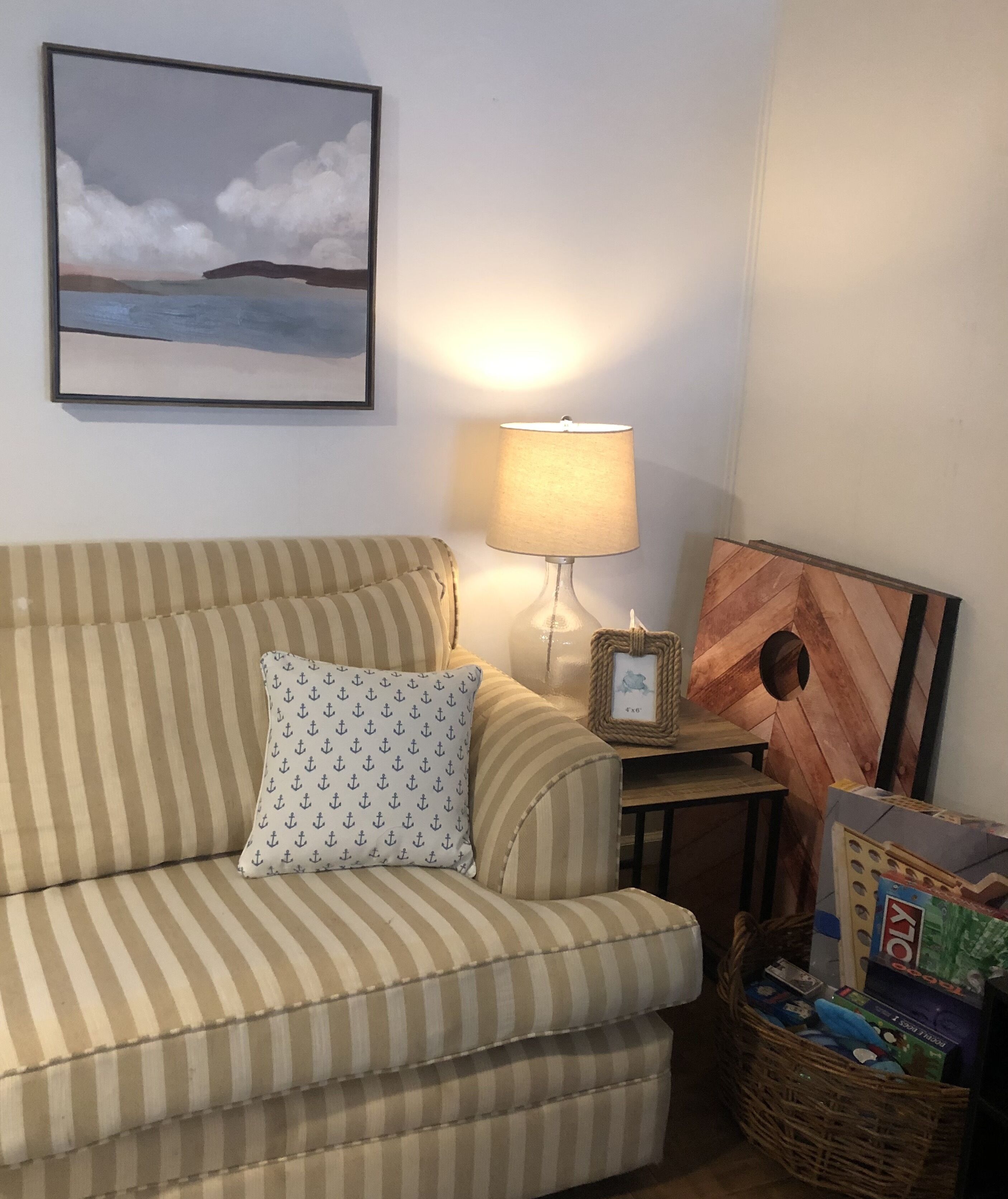 A striped sofa with a patterned pillow, a table with a lamp, a framed picture, and a basket of items.