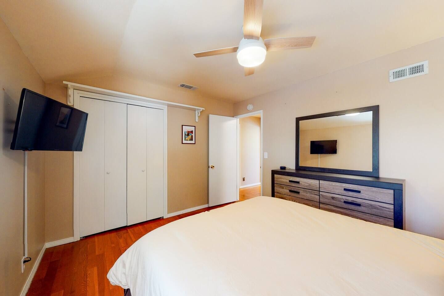 Une chambre à coucher comprenant un lit, un téléviseur fixé au mur, un ventilateur de plafond et une commode avec un miroir.