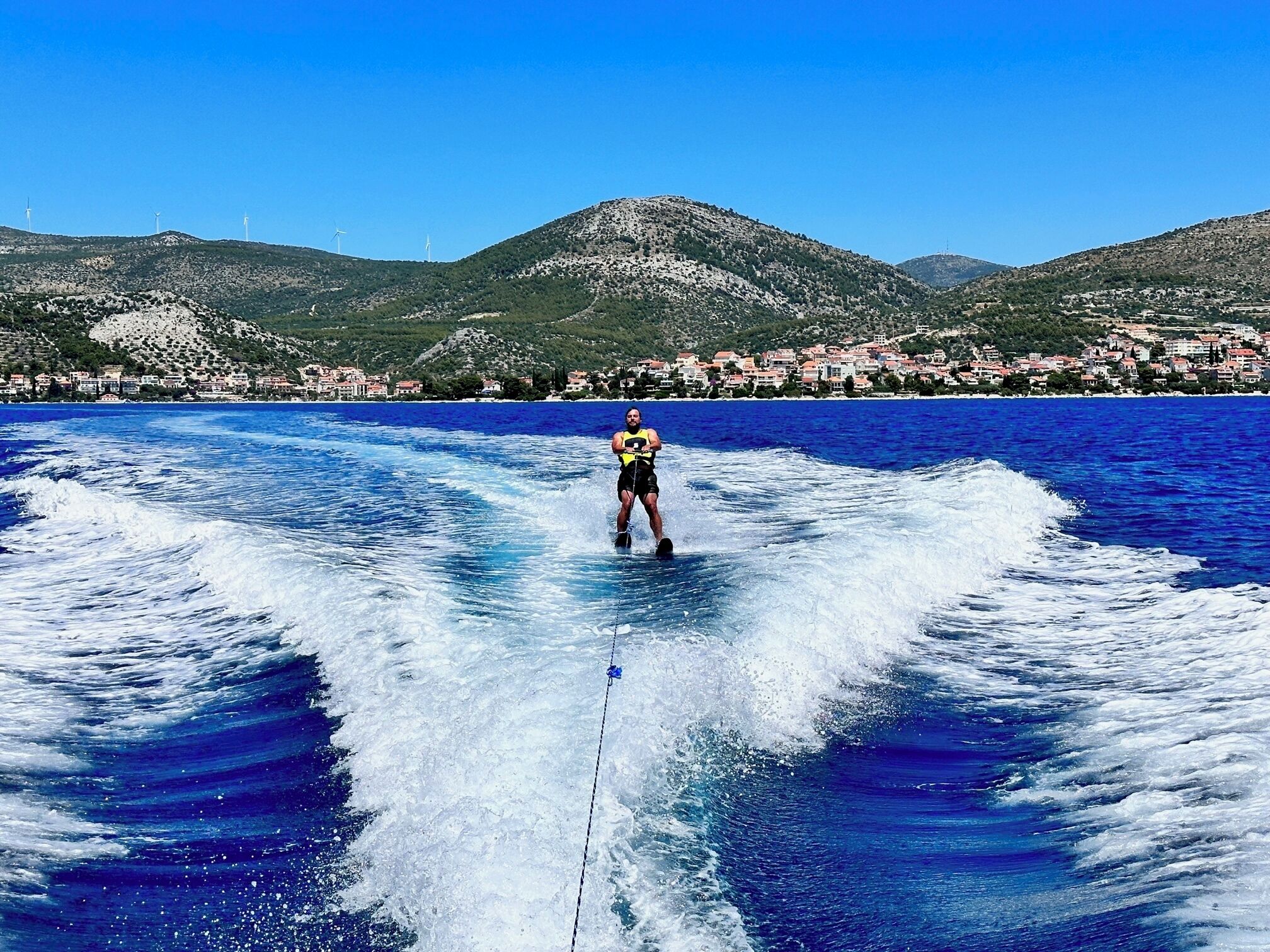 A person wakeboarding on a lake with a mountainous backdrop and a coastal town.