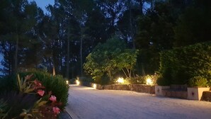 A gravel driveway leading to a stone wall with illuminated lights, surrounded by dense trees and flowering plants. - 2 apartments in a beautiful, quiet property between vineyards and the sea. Large swimming pool (LES ARCS)