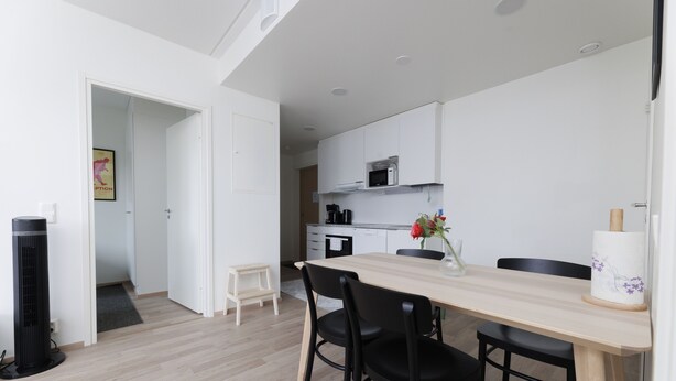 A modern kitchen with white cabinets, a microwave, and a small dining area with a wooden table and black chairs. - Hiisi Homes Hämeenlinna Asemanranta (Hameenlinna)