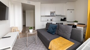 A modern living room with a grey sofa, a small coffee table, and a TV on a white stand. A kitchen with white cabinets and a microwave is visible in the background. - Hiisi Homes Hämeenlinna Asemanranta (Hameenlinna)