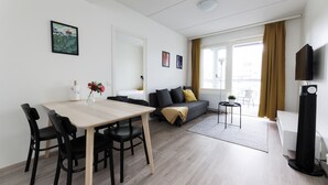 A modern living room with a dining table, black chairs, a grey sofa with yellow cushions, a small side table, and a TV on a stand. - Hiisi Homes Hämeenlinna Asemanranta (Hameenlinna)