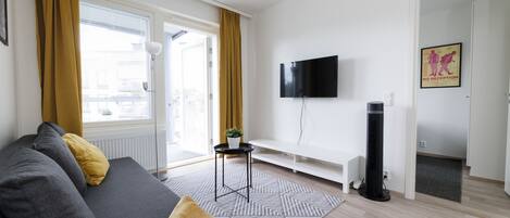 A modern living room with a grey sofa, a small side table with a plant, a black standing fan, and a mounted TV.