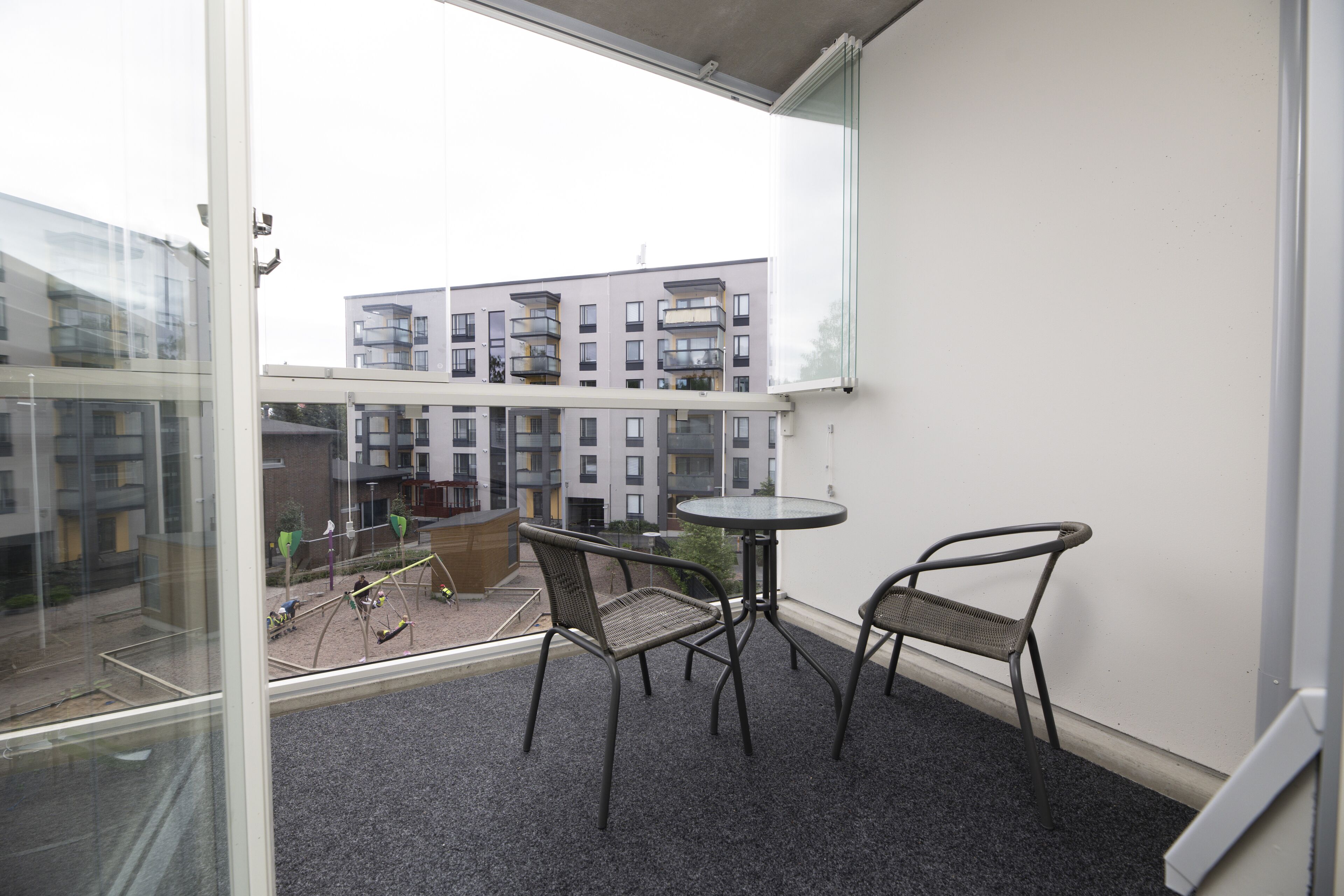 A balcony with two chairs and a small table, overlooking a courtyard with buildings and a playground.