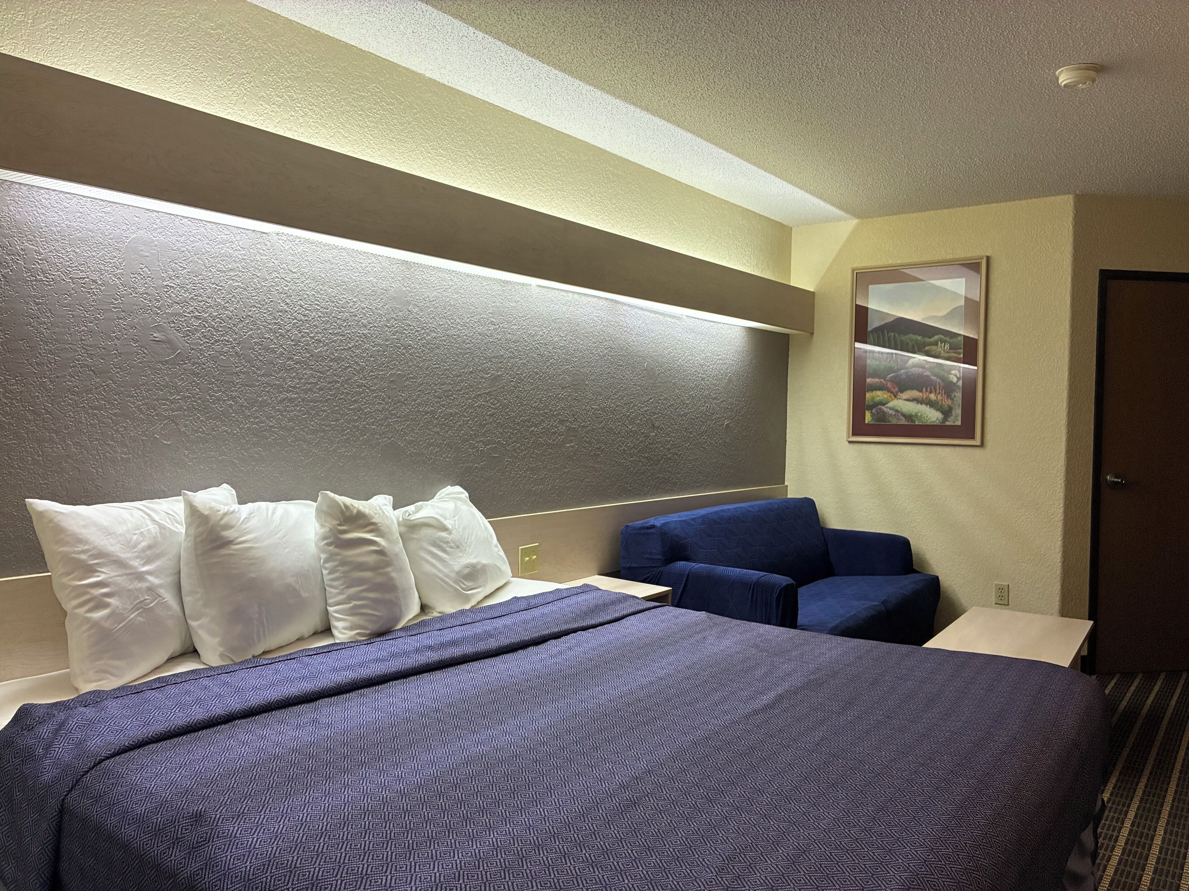 A hotel room with a bed, pillows, a blue chair, a painting on the wall, and a door.