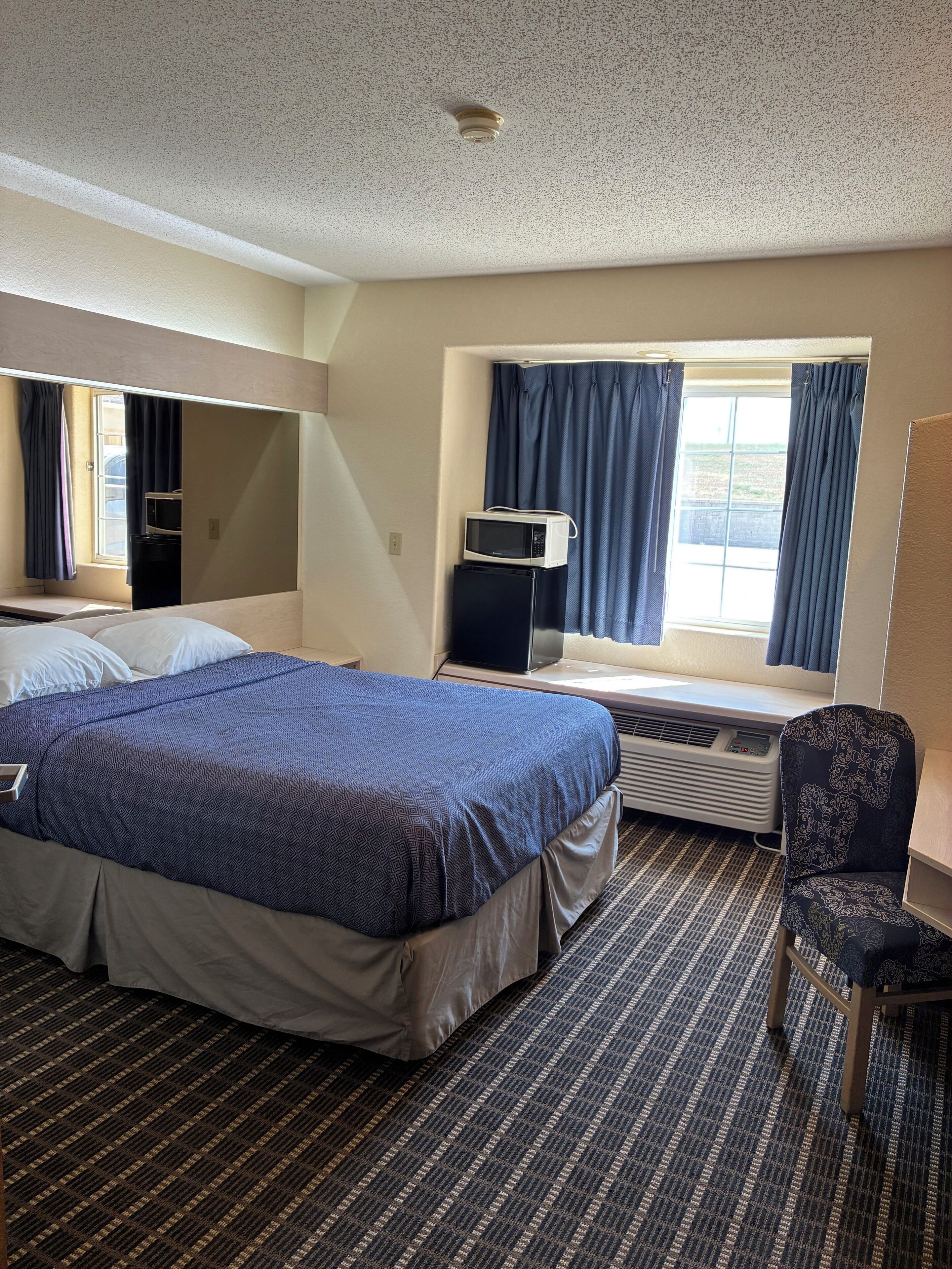 A hotel room with a large bed, a desk with a microwave, a chair, and a window with curtains.