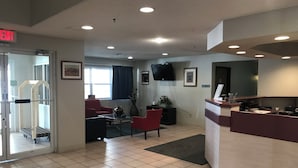 A hotel lobby with a reception desk, a TV mounted on the wall, a red chair, a coffee table, and a window with curtains.